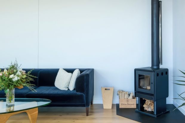 A bright, minimalist living room with a deep navy velvet sofa, pale cushions, a glass-topped coffee table, and a freestanding black wood-burning stove with flue against a white wall. Light timber flooring, a flower arrangement, firewood storage, and indoor plants add warmth and natural detail.