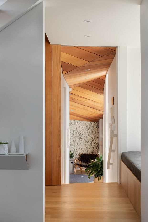 Interior view through a compact hallway or landing, featuring warm timber flooring, a sculptural timber-clad ceiling and doorway, white walls, and built-in timber cabinetry with a cushioned bench on the right. A seating area is visible beyond the opening, framed by a textured natural-stone wall and indoor plants. The design combines clean contemporary detailing with warm natural materials.