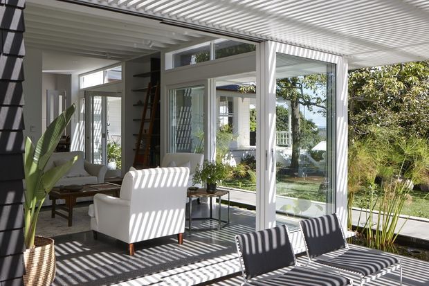 Bright glazed sunroom or enclosed outdoor living space with extensive white-framed windows and sliding doors, a white slatted pergola-style ceiling, light walls, and a dark tiled floor. White lounge chairs, a low timber coffee table, and modern outdoor chairs occupy the space, while strong linear shadows create a graphic pattern across the floor and furniture. The room opens visually to a landscaped garden and a verandah beyond.