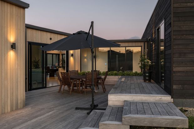 Contemporary courtyard terrace framed by low-rise timber-clad building wings. A raised grey timber deck surrounds an outdoor dining setting with a large dark umbrella, while glazed doors connect the deck to the interior. Warm wall lights, dark timber cladding, pale vertical timber boarding, and planted courtyard beds create an intimate evening outdoor space.