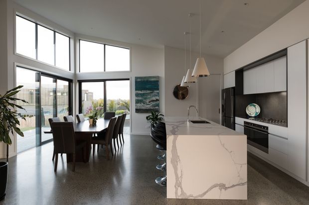 Bright contemporary open-plan kitchen and dining area with polished concrete or terrazzo-style flooring, expansive floor-to-ceiling glazing, dark dining furniture, and a large white stone island with dramatic grey veining. The kitchen features sleek white handleless cabinetry, dark splashback and integrated appliances, while pendant lights hang above the island.