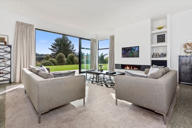 Bright contemporary living room with two large upholstered sofas arranged around a glass coffee table and patterned rug. Floor-to-ceiling glazing opens to a green garden, while a recessed fireplace, wall-mounted television, and built-in shelving form the main feature wall. The room has a clean white palette, abundant natural light, and polished concrete flooring.