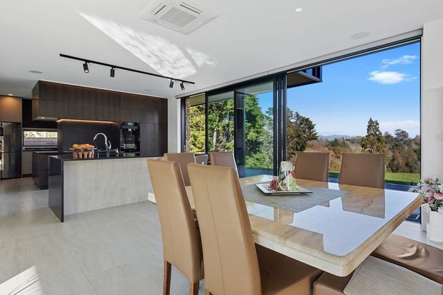 Bright open-plan kitchen and dining area with dark timber cabinetry, a pale stone island, a large polished dining table, and tan upholstered dining chairs. Full-height corner glazing opens the room to expansive views of trees, lawn, and distant hills. Contemporary track lighting and clean-lined finishes create a refined indoor-outdoor connection.