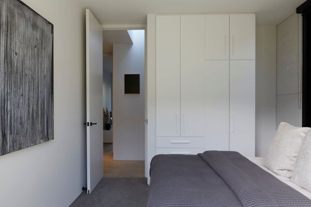 A compact, contemporary bedroom with a neatly made bed in the foreground, full-height white built-in wardrobes along the rear wall, and a white hinged door opening to a softly lit hallway. The room has pale walls, grey carpet, a textured grey bedcover, and minimal artwork, creating a clean and restrained atmosphere.