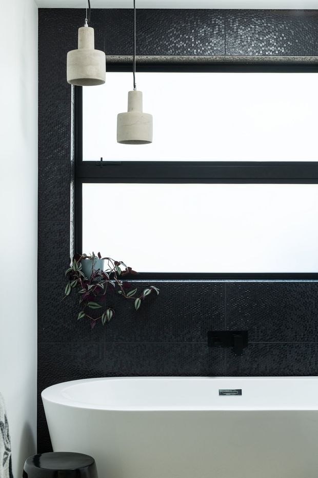 A contemporary bathroom featuring a freestanding white bathtub, black wall-mounted tapware, large black-framed windows, dark textured wall tiles, and two suspended concrete pendant lights. A trailing plant adds a small natural accent beside the bath.