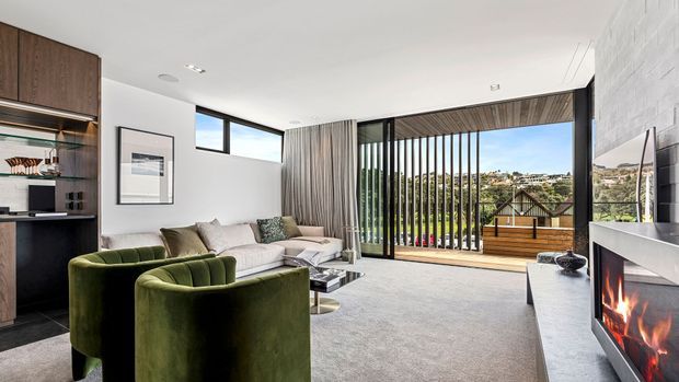 Contemporary living room with a neutral palette, green velvet armchairs, pale sectional sofa, dark timber built-in shelving, and a sleek linear fireplace. Full-height glazing with vertical timber screening opens to a timber deck and leafy suburban outlook.