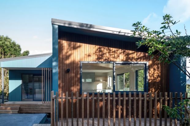 Exterior view of a compact contemporary single-storey home with a low-pitched roof, vertical timber-clad central facade, dark metal framing, and large glazed openings. A timber picket fence and deck sit in the foreground, with a glimpse of the bright interior through the open glazing.