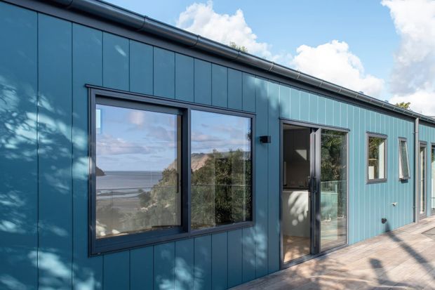 Exterior view of a contemporary single-storey building with dark blue vertical metal cladding, dark framed glazing and a glazed entry door. The facade opens onto a paved terrace, with coastal water and rocky landscape visible through the windows and door.