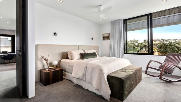 Bright contemporary bedroom with a large upholstered bed, neutral bedding, dark timber bedside table, textured bench seat, and a wooden rocking chair. Full-height glazing opens to a covered outdoor area and leafy suburban views, while a hallway or adjacent sitting area is visible through the opening on the left.