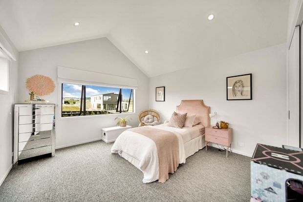 Bright, spacious bedroom with a pitched ceiling and large window overlooking an exterior view. The room features a blush pink upholstered bedhead, pale pink bedding and bedside table, white walls and cabinetry, grey carpet, framed artwork, and recessed ceiling lighting. A small desk or storage box is visible in the foreground.