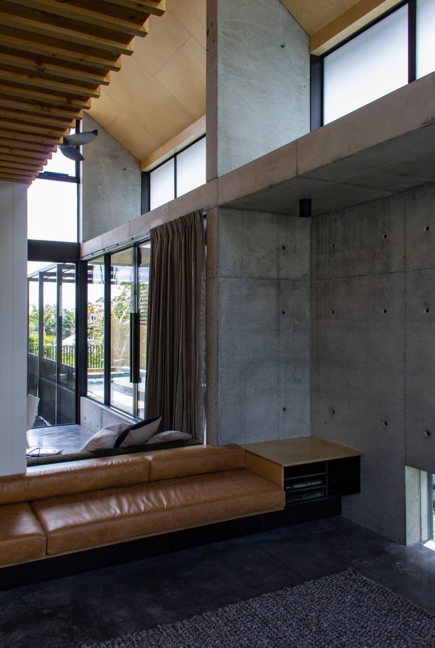 A dramatic double-height living room with exposed concrete walls, extensive black-framed glazing, and a timber-lined ceiling with exposed beams. A built-in tan leather sectional wraps around a low timber and dark joinery unit, while full-height curtains and a dark rug add warmth and softness. The space has an industrial-modern character with strong natural light and views to greenery outside.