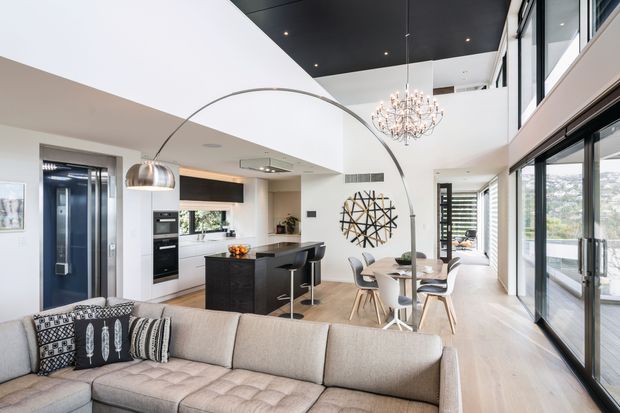 Bright open-plan contemporary living area with a large sectional sofa in the foreground, a dark kitchen island and integrated appliances, dining table, sculptural pendant lighting, double-height ceiling, and extensive floor-to-ceiling glazing opening to a balcony or exterior view. The space uses light timber flooring, white walls, black ceiling elements, and dark kitchen cabinetry.