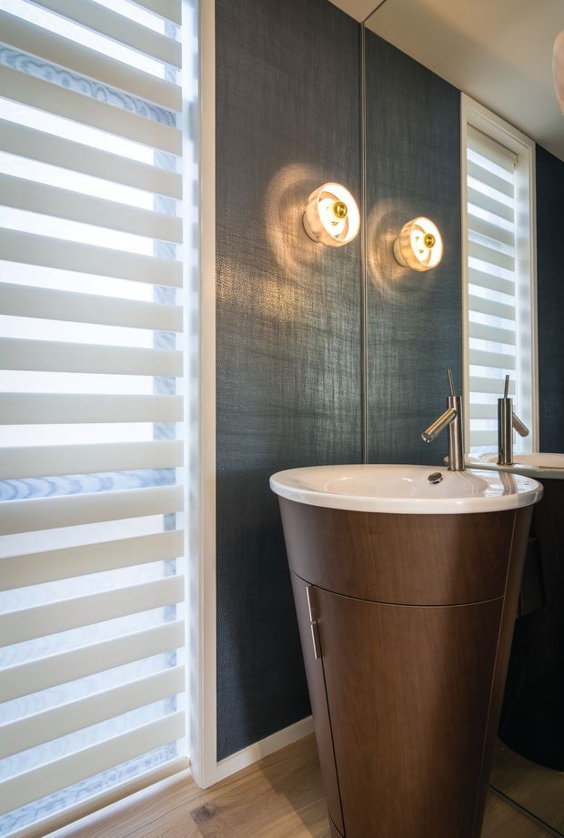 A compact, contemporary bathroom featuring a sculptural freestanding timber pedestal basin with chrome tapware, dark textured wall panelling, twin round wall lights, a large mirror, and layered white roller blinds over tall windows. Timber flooring adds warmth to the dark, refined palette.