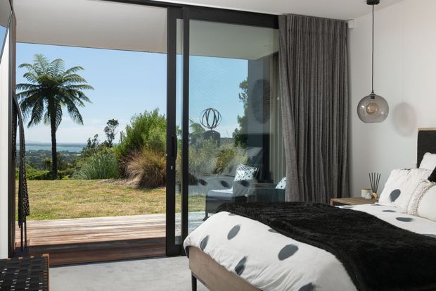 A contemporary bedroom with a low-profile upholstered bed, white bedding with black circular accents, and a dark textured throw. Floor-to-ceiling black-framed sliding glass doors open to a timber deck and landscaped coastal-looking view, while full-height grey curtains and a spherical pendant light add a refined, modern finish.