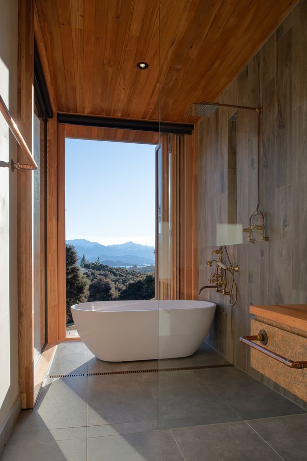 A refined contemporary bathroom with a freestanding white bathtub positioned beside full-height glazing overlooking a mountainous landscape. The room features large grey floor tiles, grey stone-look wall tiles, warm timber wall and ceiling lining, a frameless glass shower screen, and aged brass shower and tapware. A timber vanity with a stone benchtop is visible on the right.