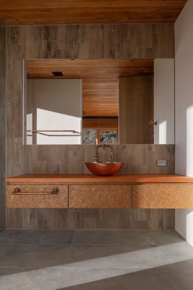 A warm, contemporary bathroom vanity with a floating textured brown timber or stone-effect cabinet, wood-toned benchtop, copper vessel basin and matching vintage-style tapware. The walls are clad in large wood-look tiles, complemented by dark grey floor tiles and a broad mirror. A timber-lined ceiling and reflected interior add warmth, while strong sunlight creates diagonal shadows across the floor.