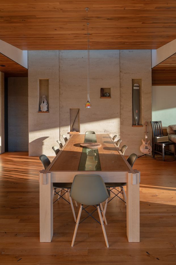 A warm, sunlit dining area centered on a long rectangular timber dining table with a glass inset and multiple modern chairs. The room features extensive timber flooring and a timber-lined ceiling, a large exposed concrete feature wall with recessed display niches, a slim pendant light, and an adjacent living area with a guitar and seating visible at right.