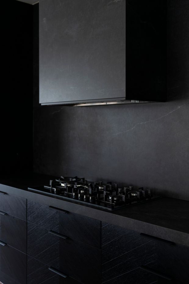 A dark, monochromatic contemporary kitchen vignette featuring black textured cabinetry with integrated drawers, a black gas cooktop, dark stone-look benchtop and matching full-height splashback, plus a large black rangehood. The space is dramatically lit with a moody, refined aesthetic.