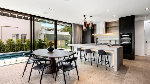 Contemporary open-plan kitchen and dining area with a round dark timber dining table and black chairs beside full-height glazing. The kitchen features a large light stone island with four bar stools, dark integrated cabinetry, timber-look wall cabinetry, pendant lights, and tiled flooring. A landscaped courtyard and pool are visible outside.