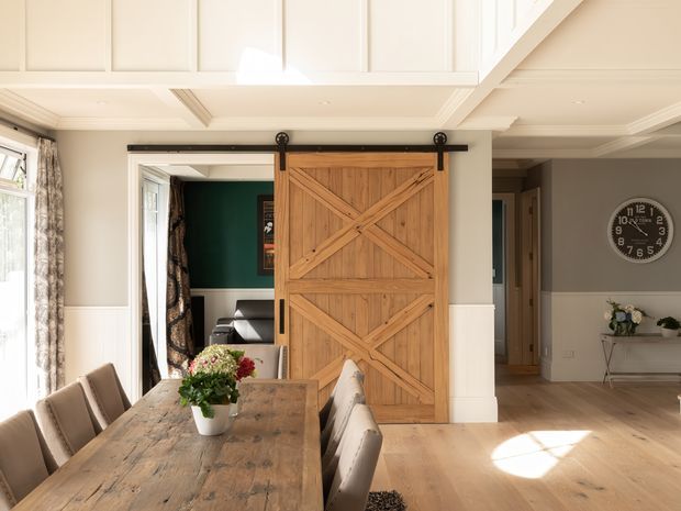 Bright, refined dining and living area featuring a large rustic timber dining table, upholstered dining chairs, white wall panelling and coffered ceiling detailing. A prominent sliding barn door with diagonal bracing separates the dining area from a dark green adjoining room, while a hallway and simple console styling are visible beyond. Natural timber flooring and warm daylight create a welcoming transitional interior.