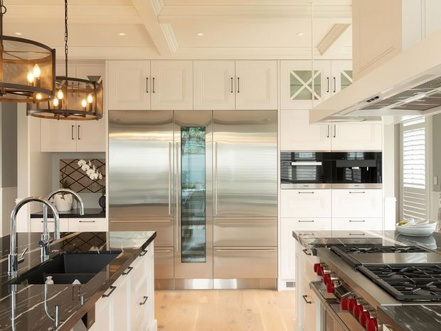 Bright, spacious luxury kitchen with extensive white built-in cabinetry, stainless-steel double-door refrigeration, black-and-white veined stone benchtops, integrated appliances, a large gas cooktop, and twin islands or work zones. The room features a coffered white ceiling, polished metal hardware, dark pendant lighting, and pale timber flooring.