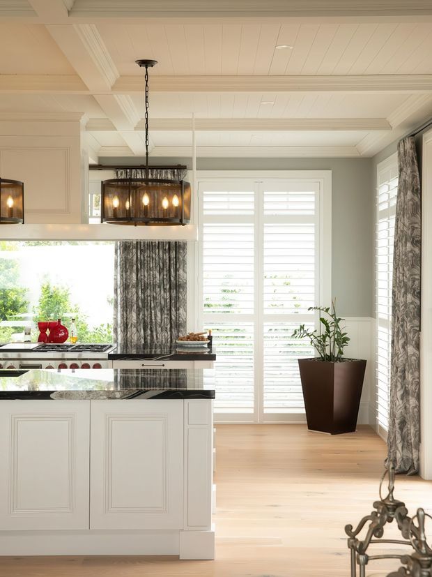 Bright, refined kitchen interior with a large white paneled island, dark stone benchtops, white cabinetry, timber flooring, plantation shutters, patterned curtains, and a prominent rectangular pendant light. The room has a light traditional architectural character with decorative ceiling beams and detailed trim, opening toward a sunlit glazed area.