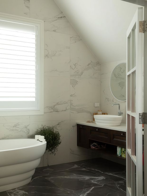 A compact, bright bathroom with dramatic marble-look wall and floor tiles. The room features a freestanding white bathtub beneath a shuttered window, a floating dark timber vanity with a white vessel basin and chrome tapware, a round mirror, and a partially open glazed-panel door. The sloped ceiling creates an attic-like configuration.