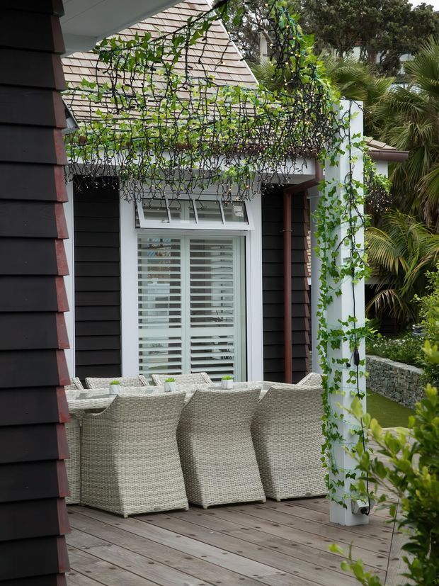A covered outdoor dining terrace beside a dark-clad home, with a timber deck, woven outdoor dining chairs and table, white-framed louvred window, and a white pergola structure heavily covered in climbing vines. Tropical planting and a low stone retaining wall surround the space, creating a sheltered garden setting.