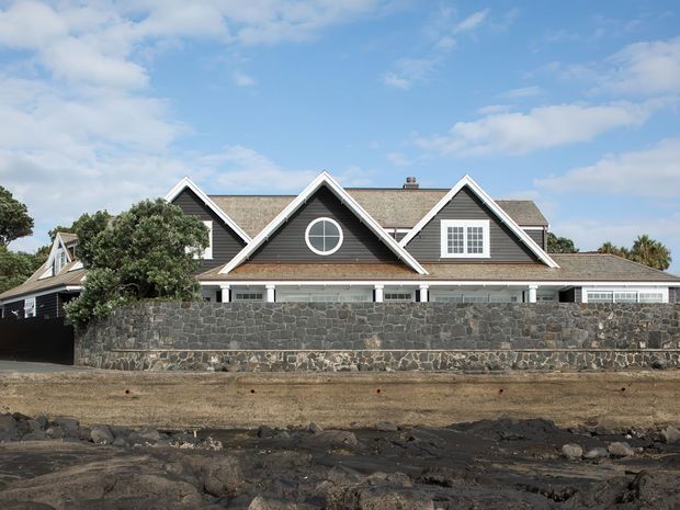 Exterior view of a large, low-rise residential house behind a dark volcanic-stone boundary wall. The home has multiple steep front-facing gables, grey shingle-like roof surfaces, white trim, dormer windows, and a circular feature window. It is set in a stark rocky landscape under a bright partly cloudy sky.