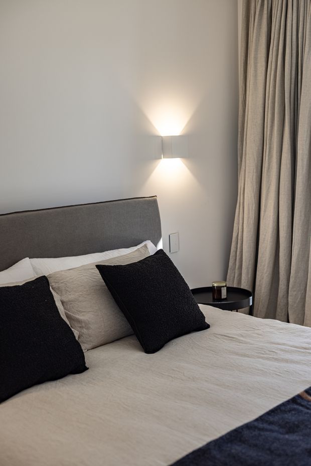 A softly lit contemporary bedroom corner featuring a textured grey upholstered headboard, layered white and beige bedding, black accent cushions, and a small round black bedside table. A sculptural wall sconce casts an upward and downward glow on the pale wall, while full-height taupe curtains frame the right side.