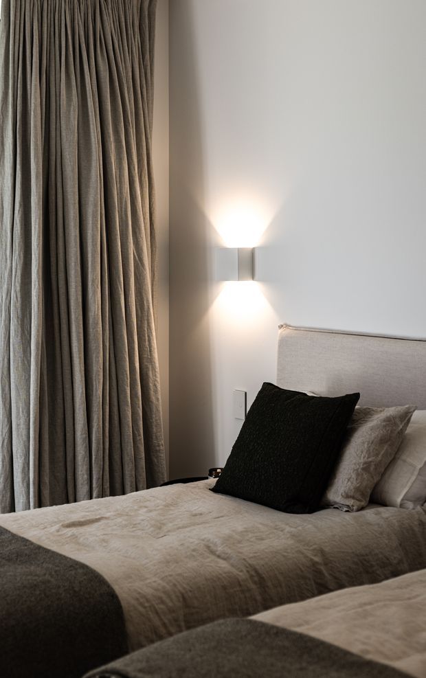 A softly lit bedroom corner featuring a neatly made bed with layered beige and taupe bedding, a black accent cushion, and a light upholstered headboard. Full-height pleated taupe curtains frame the left side, while a minimalist wall sconce casts warm up-and-down light on the pale wall.