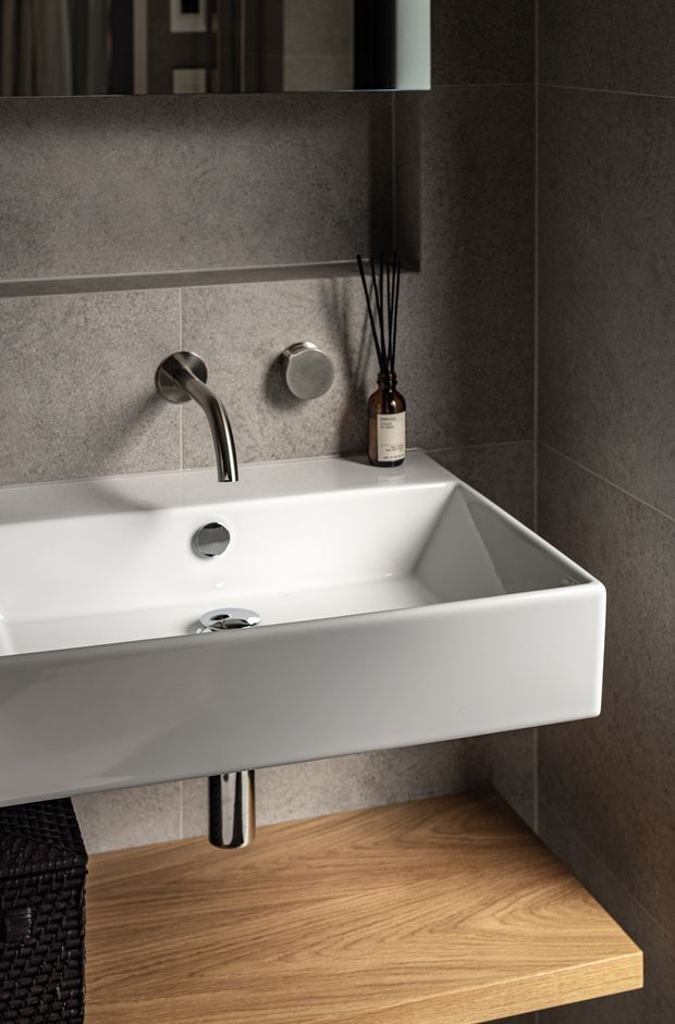 Close-up view of a contemporary bathroom vanity featuring a wall-mounted white rectangular basin, chrome wall-mounted tapware, grey large-format tiled walls, a frameless mirror, and a light timber floating shelf beneath the basin. A small reed diffuser adds minimal styling.