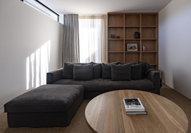 A contemporary living room with a large dark charcoal L-shaped sectional sofa and ottoman, a round light timber coffee table, and a full-height built-in timber shelving unit displaying a few books and decorative objects. Sheer curtains and grey drapes cover a tall window, with strong daylight casting linear shadows across the pale walls. The room has a restrained, warm-neutral palette and a clean, relaxed design.
