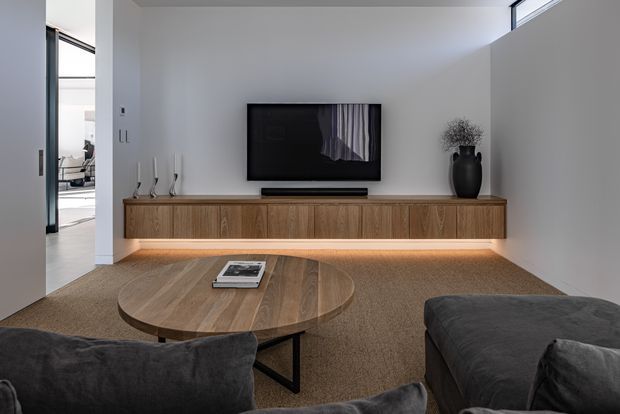 A refined contemporary living room with a wall-mounted television above a long floating timber entertainment console. The room features warm wood tones, a rounded timber coffee table, dark upholstered sofas, neutral walls, recessed LED lighting beneath the joinery, and a high horizontal clerestory window. An open doorway reveals part of an adjoining space.