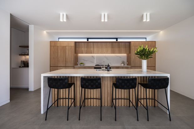 A bright contemporary kitchen viewed from the living area, centered on a large island with four black bar stools. The kitchen features warm timber cabinetry, a pale stone benchtop, a light marble-look splashback, dark flooring, integrated open shelving, and concealed linear lighting. Minimal white walls and ceiling-mounted linear fixtures create a clean, refined appearance.
