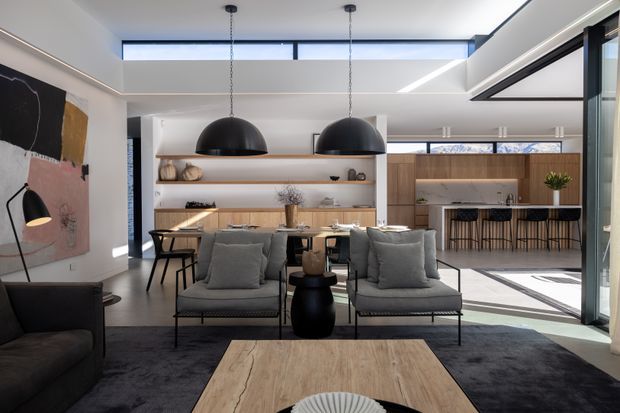 A refined open-plan living, dining and kitchen interior with a neutral palette and abundant natural light. The living area features grey upholstered lounge chairs and a dark sofa around a pale timber coffee table, with a large dark rug beneath. Behind it, a timber dining table sits beneath two oversized black dome pendant lights, while open timber shelving displays ceramics. The contemporary kitchen has warm timber cabinetry, a light benchtop, a long island with dark vertical detailing, and expansive glazing. A large skylight and linear ceiling lighting enhance the airy, high-quality interior.