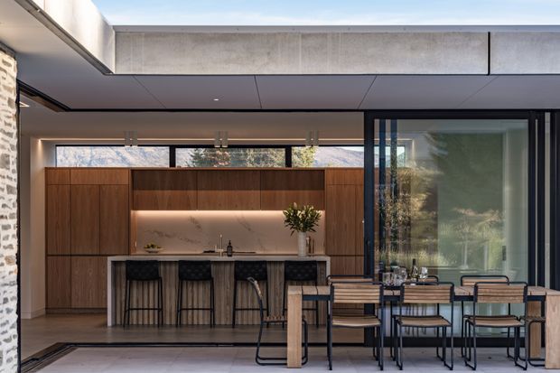A refined contemporary open-plan kitchen and dining area opening to the outdoors. The kitchen features dark timber cabinetry, a softly illuminated stone-look splashback, a long island with bar seating, and integrated warm lighting. A timber dining table with modern black-framed chairs occupies the foreground, while extensive glazing provides views of trees and mountains. The space is sheltered beneath a flat roof with exposed concrete fascia and dark ceiling panels.