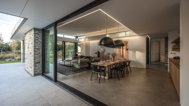 Open-plan contemporary living, dining and kitchen area viewed from a covered outdoor terrace. Full-height black-framed glazing and sliding doors connect the interior to the landscaped exterior. The room features a large dining table, upholstered seating, pendant lighting, timber kitchen cabinetry, a concrete-look floor, and a prominent exposed brick-clad structural wall.