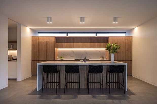 A contemporary kitchen with a large central island and four black bar stools. Full-height warm timber cabinetry spans the back wall, incorporating open shelving and a softly illuminated stone-look splashback. Integrated appliances and a sink are positioned along the rear bench, while recessed ceiling lights and linear clerestory windows provide a clean, softly lit atmosphere. The island has a pale stone benchtop and dark vertical-panel detailing.