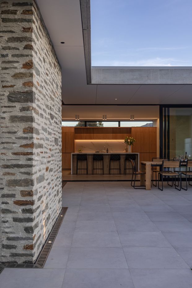A refined open-plan indoor-outdoor living and entertaining area viewed beneath a covered roof structure. A substantial exposed masonry wall of irregular pale stone with darker and warm brown inclusions dominates the foreground. Beyond it is a contemporary kitchen with warm timber cabinetry, a stone island benchtop, integrated lighting and dark bar stools. A dining table and chairs sit to the right beside large glazed openings. Large-format pale grey floor tiles continue across the space, while a broad roof opening frames the sky and adds strong architectural interest.