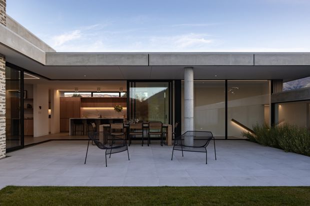 Contemporary single-level residence viewed from a spacious paved terrace. The home has a broad flat roof with deep overhangs, extensive floor-to-ceiling dark-framed glazing, a cylindrical concrete column, and a partially exposed brick wall. Through the open glazing is a warm, timber-lined kitchen and dining area with a long island, while minimalist outdoor dining and lounge furniture occupies the terrace. Low ornamental planting borders the facade.