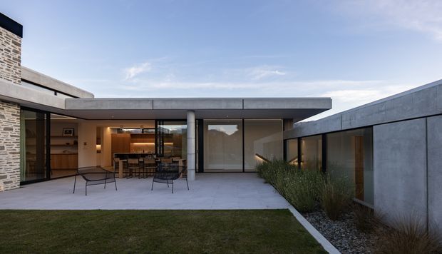 Exterior view of a contemporary single-storey residence arranged around a paved courtyard. The composition features a low flat roof with broad concrete fascia, extensive floor-to-ceiling glazing, a stone-clad wall at left, and a projecting concrete wing at right. Through the open glazing, a warm timber-lined kitchen or dining area is visible. The courtyard has minimalist paving, restrained planting, and a small lawn.
