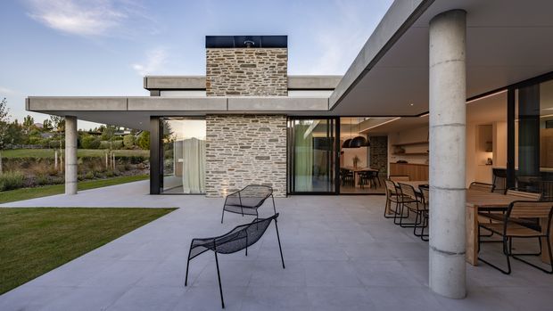 Contemporary single-storey residence viewed from a covered outdoor terrace, featuring broad flat roof planes, exposed cylindrical concrete columns, extensive glazing, and a central rough stone-clad chimney. The terrace is furnished with modern outdoor seating and dining furniture, with the open-plan interior visible beyond the glass doors. Landscaped lawn and garden surround the house.