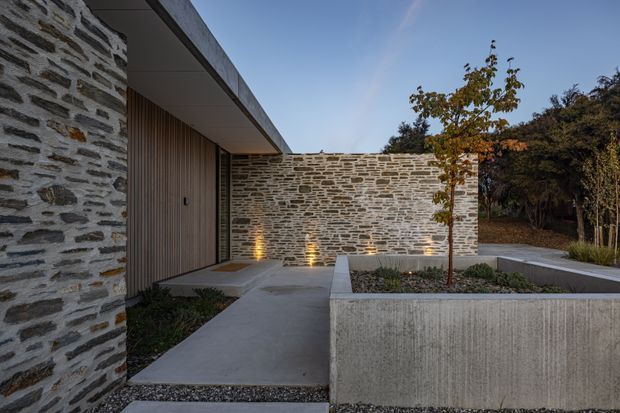 A contemporary low-profile building exterior arranged around a landscaped courtyard. Rough layered stone walls contrast with smooth concrete paving and raised planters, while vertical timber-clad entry doors sit beneath a broad flat roof overhang. Warm recessed uplighting highlights the stonework, and a young tree and garden planting add a restrained natural element.