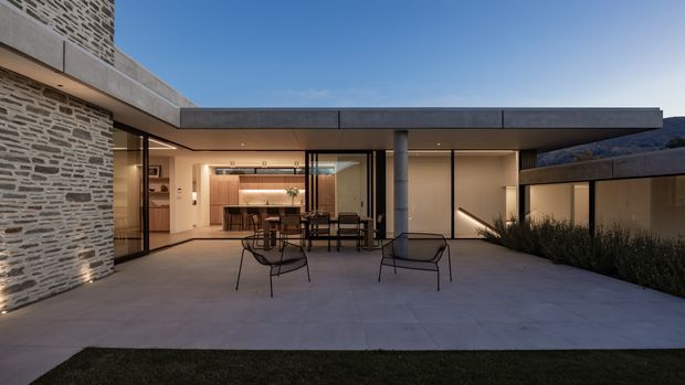 Contemporary single-storey residence opening onto a broad paved courtyard or terrace at dusk. The composition highlights a low flat roof with deep concrete overhangs, exposed cylindrical concrete columns, extensive dark-framed glazing, and a prominent textured stacked-stone wall. Through the glazing, a warm timber kitchen and dining area are visible. Sparse outdoor furniture and integrated planting create a refined indoor-outdoor setting, with a dark mountainous form visible in the background.