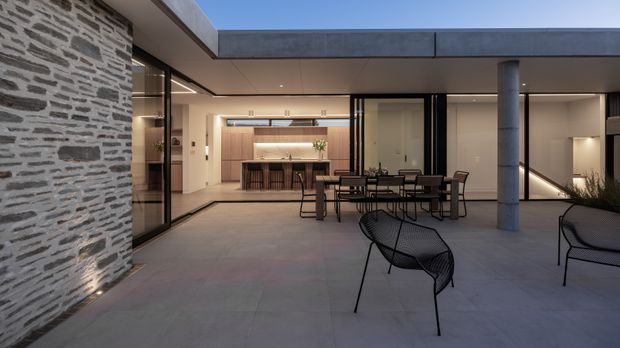 A refined contemporary courtyard terrace viewed from outdoors, with a broad flat-roofed structure, exposed grey stone feature wall, concrete column, glazed openings, and a warm illuminated kitchen and dining area beyond. The paved courtyard contains a dining table and modern wire-frame lounge chairs, creating an indoor-outdoor entertaining space.