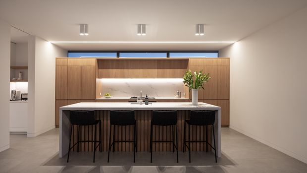 A contemporary kitchen viewed straight on, dominated by a large island with four black bar stools. The island has a dark stone-like benchtop and timber slatted cabinetry, while the rear wall features full-height warm timber cabinetry, a softly illuminated pale stone or tiled splashback, and a concealed work area. The room has a restrained neutral palette, recessed ceiling spotlights, linear LED lighting, pale flooring, and a narrow horizontal window above the cabinetry.