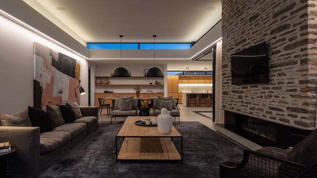 A sophisticated open-plan living room with a dark sectional sofa, central timber coffee table, lounge chairs, and a large textured rug. A dramatic exposed masonry feature wall incorporates a fireplace and wall-mounted television. The space opens to a dining area and kitchen at the rear, with warm timber shelving and cabinetry, integrated linear lighting, pendant lights, concealed strip lighting, and clerestory glazing creating a refined contemporary atmosphere.