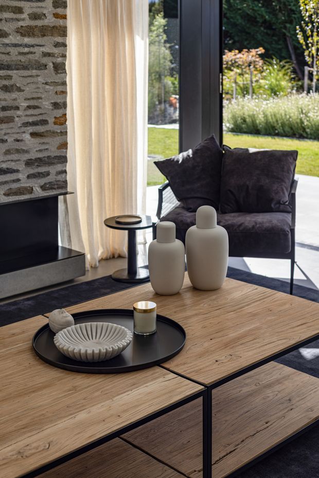 A refined contemporary living room with a low modular timber coffee table in the foreground, styled with a black tray, ceramic vessels, and a candle. A dark upholstered lounge chair sits beside a floor-to-ceiling glazed opening, with sheer cream curtains and a landscaped garden visible beyond. A rough natural stone fireplace wall and dark hearth anchor the left side of the room.