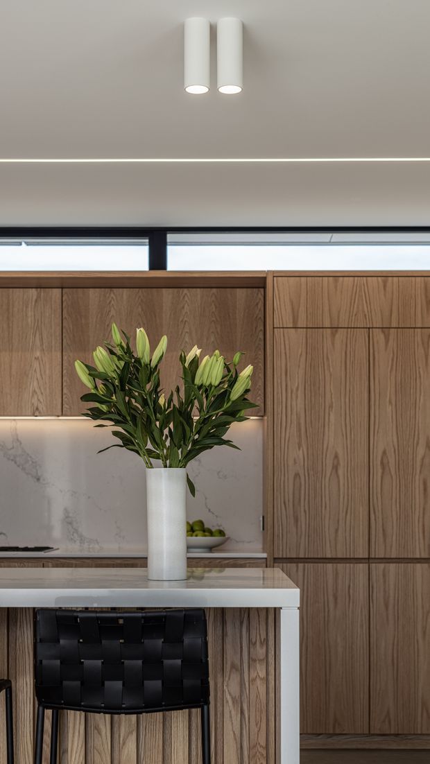 A refined contemporary kitchen detail featuring warm vertical-grain timber cabinetry, a pale veined stone splashback, and a light stone island benchtop. A tall white vase of lilies sits on the island beside a black woven bar stool. Integrated linear lighting, cylindrical white ceiling lights, and a narrow clerestory window contribute to the clean, understated composition.